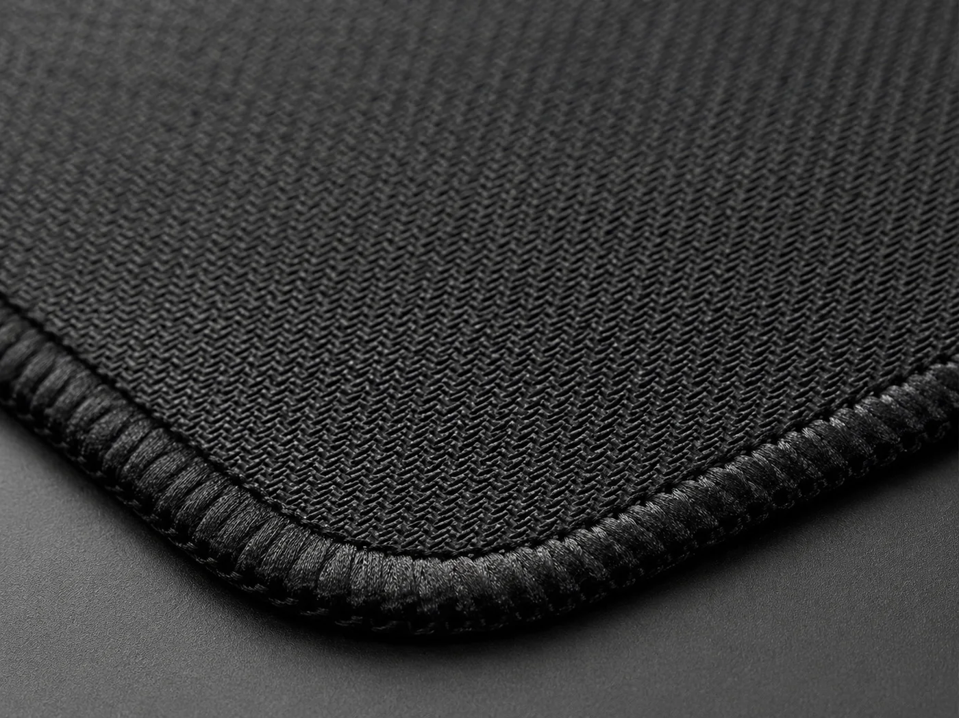 Macro close-up of a black desk mat corner on a dark surface, showing the fine fabric weave and the tight stitched edge running around the rounded corner