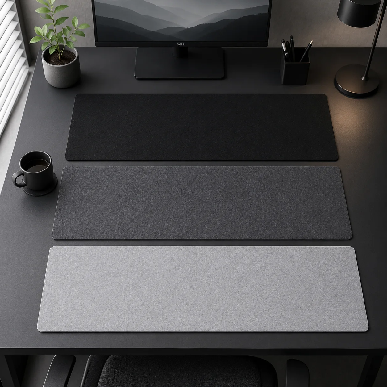 Top-down view of three plain desk mats laid one above the other on a dark desk — black, concrete gray and light gray — next to a monitor, a coffee mug and a small plant
